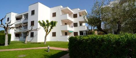 A modern white apartment building with multiple balconies, a paved pathway, and well-maintained greenery.