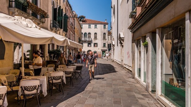 Une rue étroite avec des tables en terrasse, des bâtiments de chaque côté et un ciel dégagé.