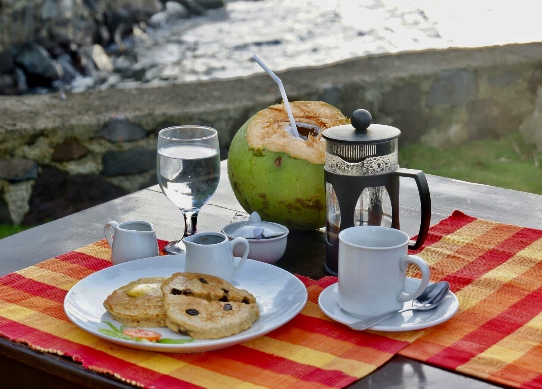 Un tavolo apparecchiato con un piatto di biscotti, un bicchiere d'acqua, una caffettiera e una tazza di caffè, insieme a un cocco e una tovaglia a quadri.