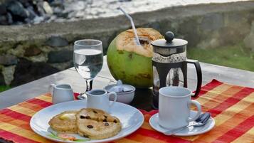 Un tavolo apparecchiato con un piatto di biscotti, un bicchiere d'acqua, una caffettiera e una tazza di caffè, insieme a un cocco e una tovaglia a quadri.