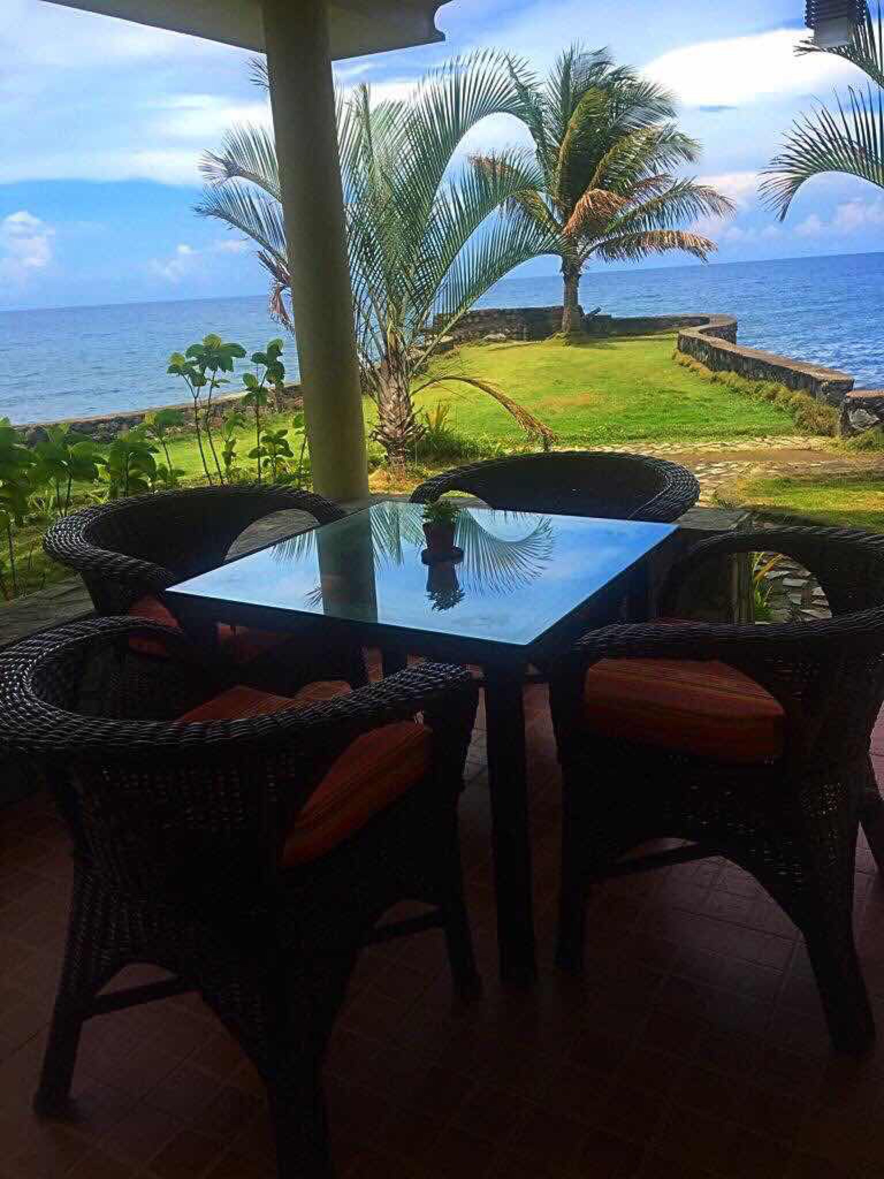 Uma mesa com tampo de vidro, cadeiras de vime e almofadas, em um pátio com vista para o mar e palmeiras.