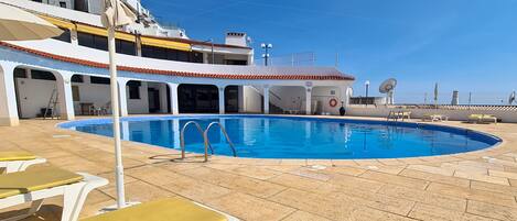 Un área de piscina con tumbonas y un flotador salvavidas, rodeada por un edificio con tejado de azulejos y antenas parabólicas.