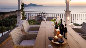 Una mesa de madera con sillas de mimbre, botellas de vino, copas y velas en un balcón con vistas al mar.