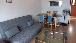 A living room with a grey sofa, a glass coffee table, wooden chairs, a small table, a standing fan, and a framed picture on the wall. - RIELLS PLATJA (Escala)