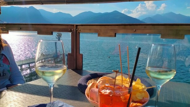 A table with two glasses of white wine, a bowl of fruit, and a drink with a straw, set against a backdrop of a lake and mountains. - FeWo Luino Loft: Historic Charm, 100m to Lake & Swiss Gateway. (Luino)