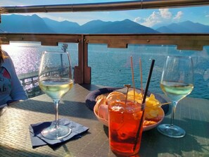 A table with two glasses of white wine, a bowl of fruit, and a drink with a straw, set against a backdrop of a lake and mountains.