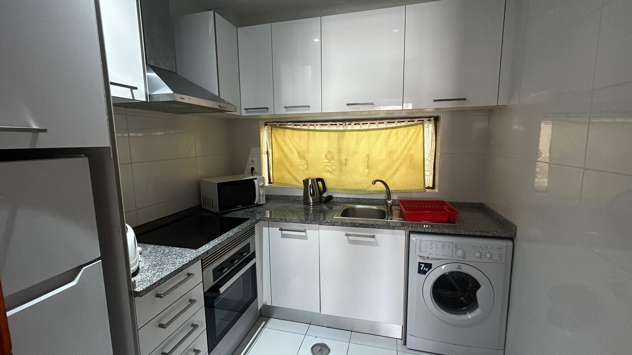 A compact kitchen with white cabinets, a microwave, a kettle, a sink, and a washing machine.