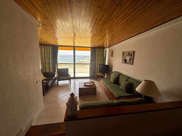 A living room with a wooden ceiling, a green sofa, a television, and a view of the sea through a glass door. - Albufeira, Ocean terrace (22) by Rentals in Algarve (Albufeira)