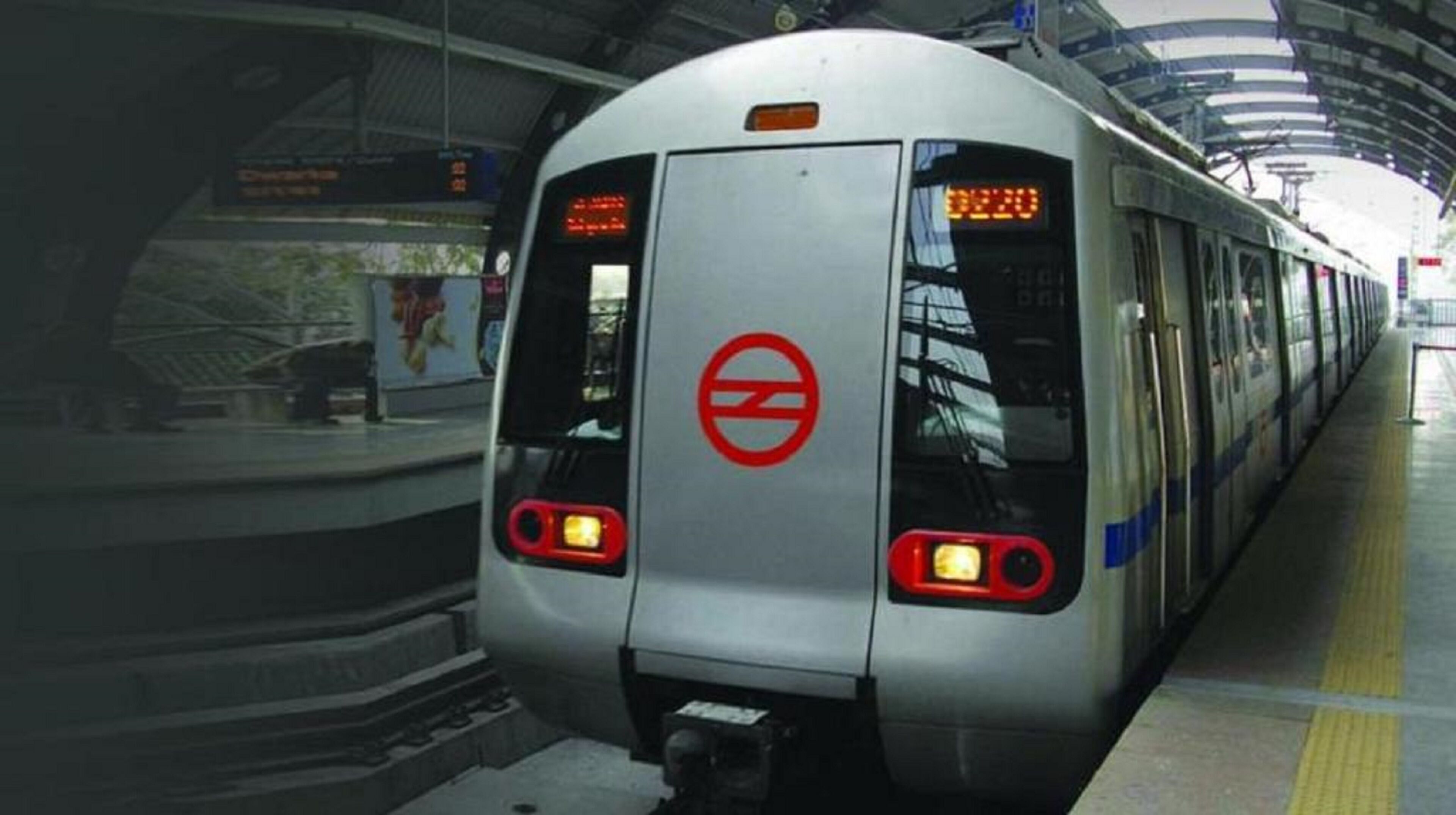 A metro train at a station with a circular logo on the back, platform edge, and overhead structure.