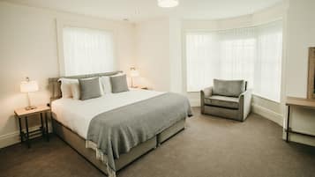 Quarto de hotel com uma cama grande, uma cadeira, uma escrivaninha e uma janela com cortinas.