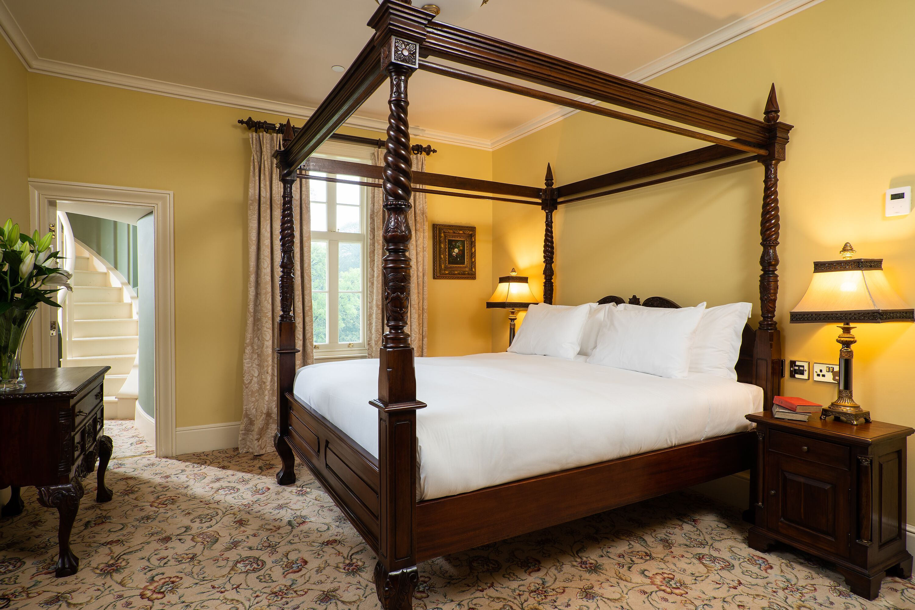 A four-poster bed with a wooden frame, a nightstand with a lamp, and a floral-patterned carpet.