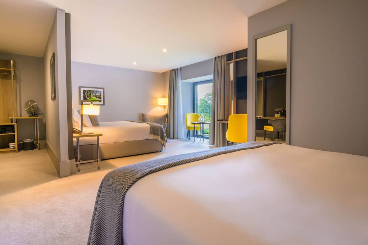 A hotel room with a large bed, a sofa, a desk, and a chair. There is a window with curtains, a mirror, and a picture on the wall.