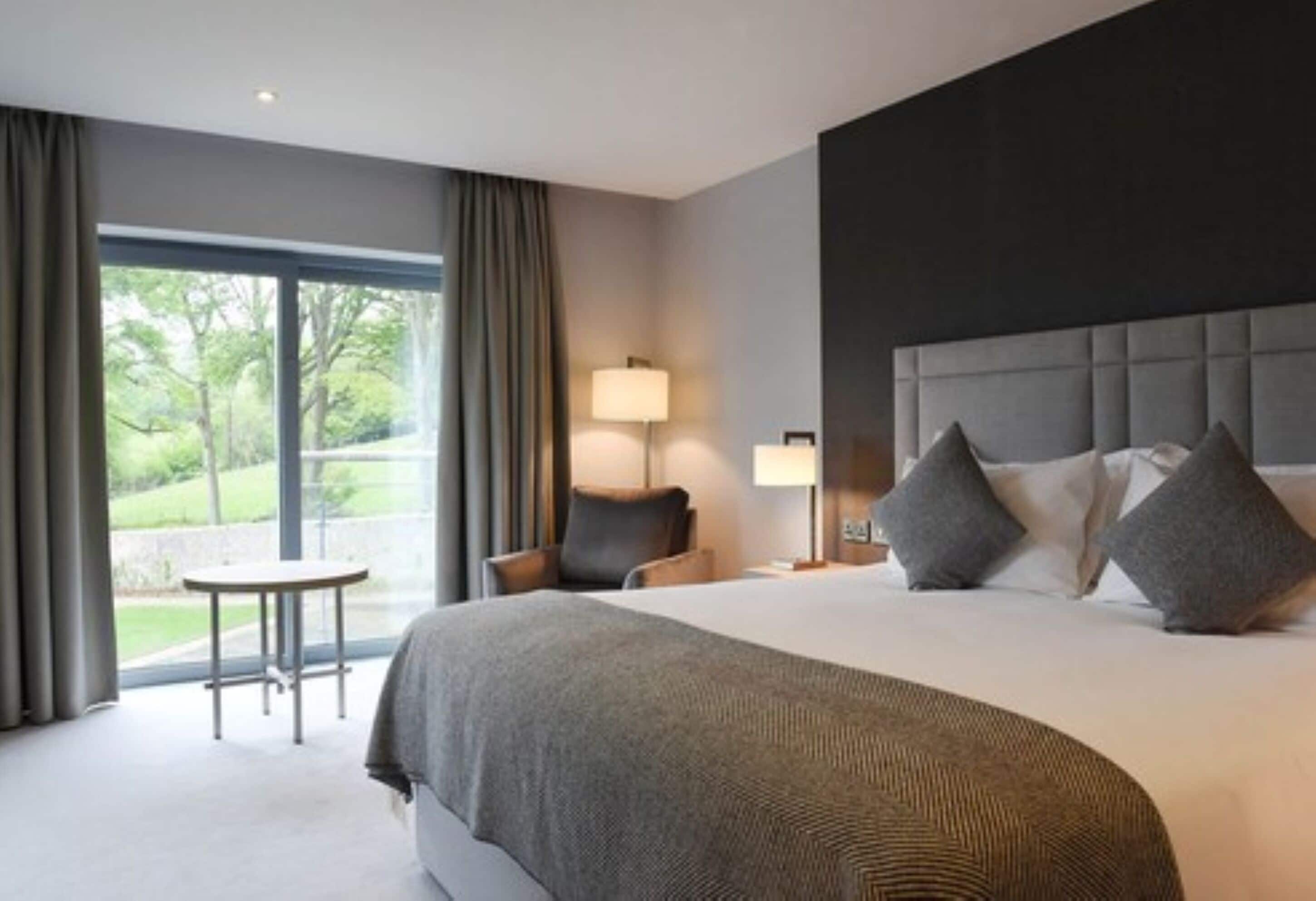 A hotel room with a large bed, a bedside table, a chair, a lamp, and a view of the outdoors.