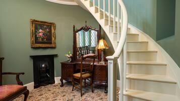 A room with a wooden desk, chair, and a fireplace. There is a staircase with white railings and a chandelier.