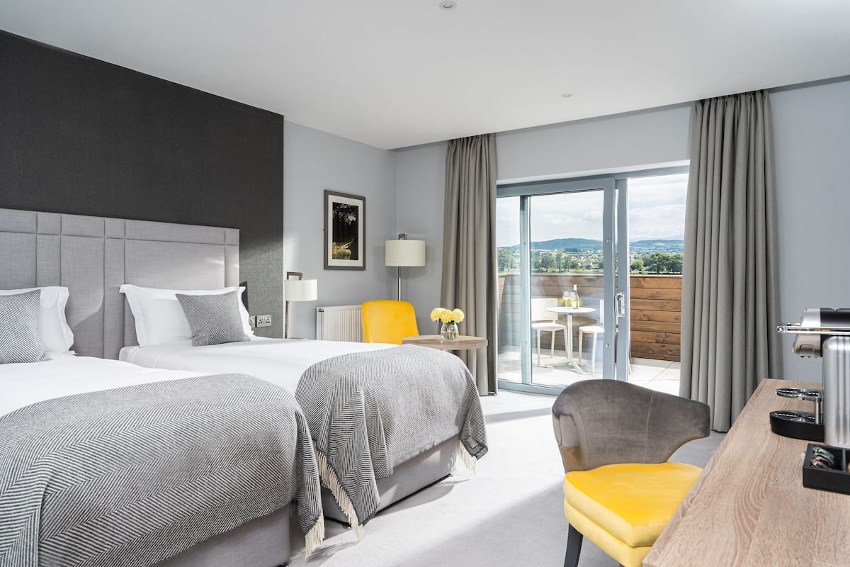 A hotel room with two beds, a desk, a chair, a lamp, and a view of the outdoors.