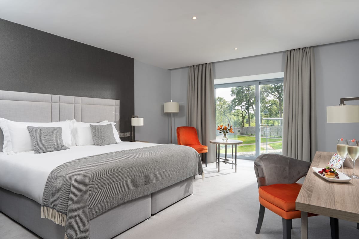 A modern hotel room with a large bed, a dining table with two chairs, and a view of the outdoors.