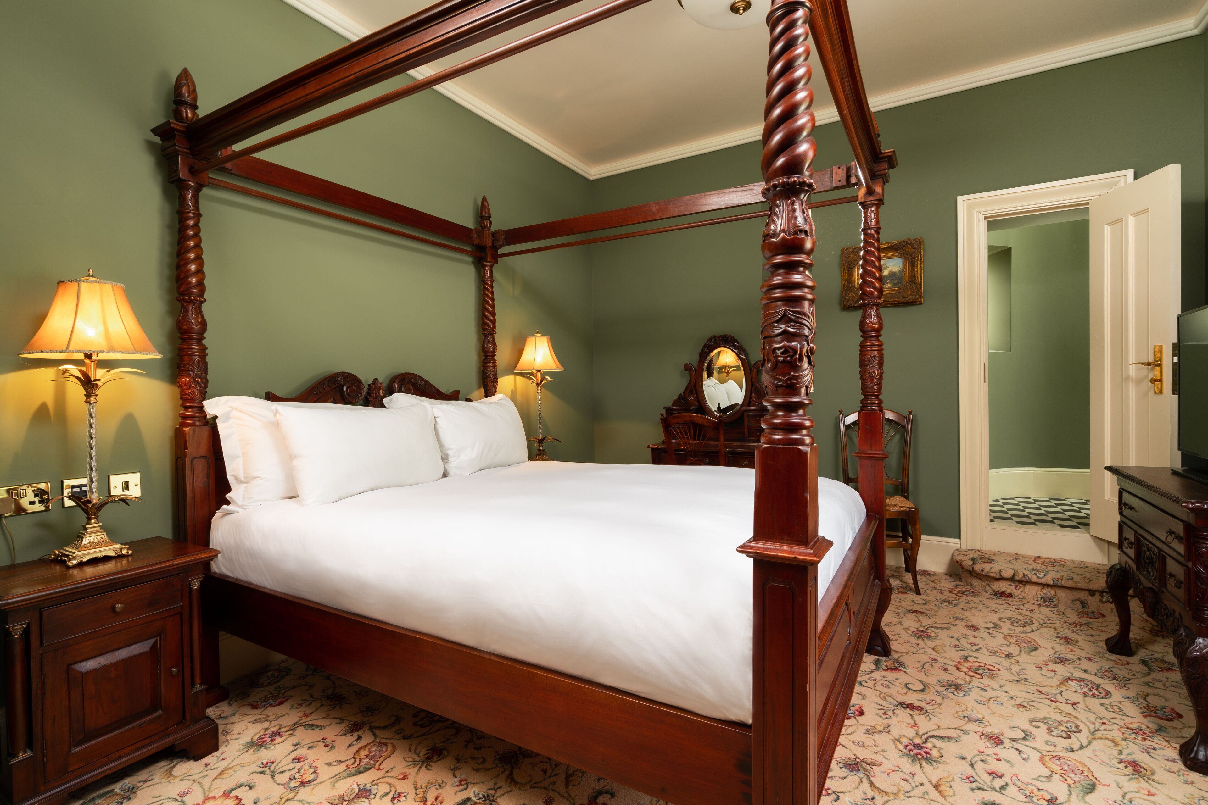 A four-poster bed with white linens, wooden nightstands with lamps, and a floral-patterned carpet.