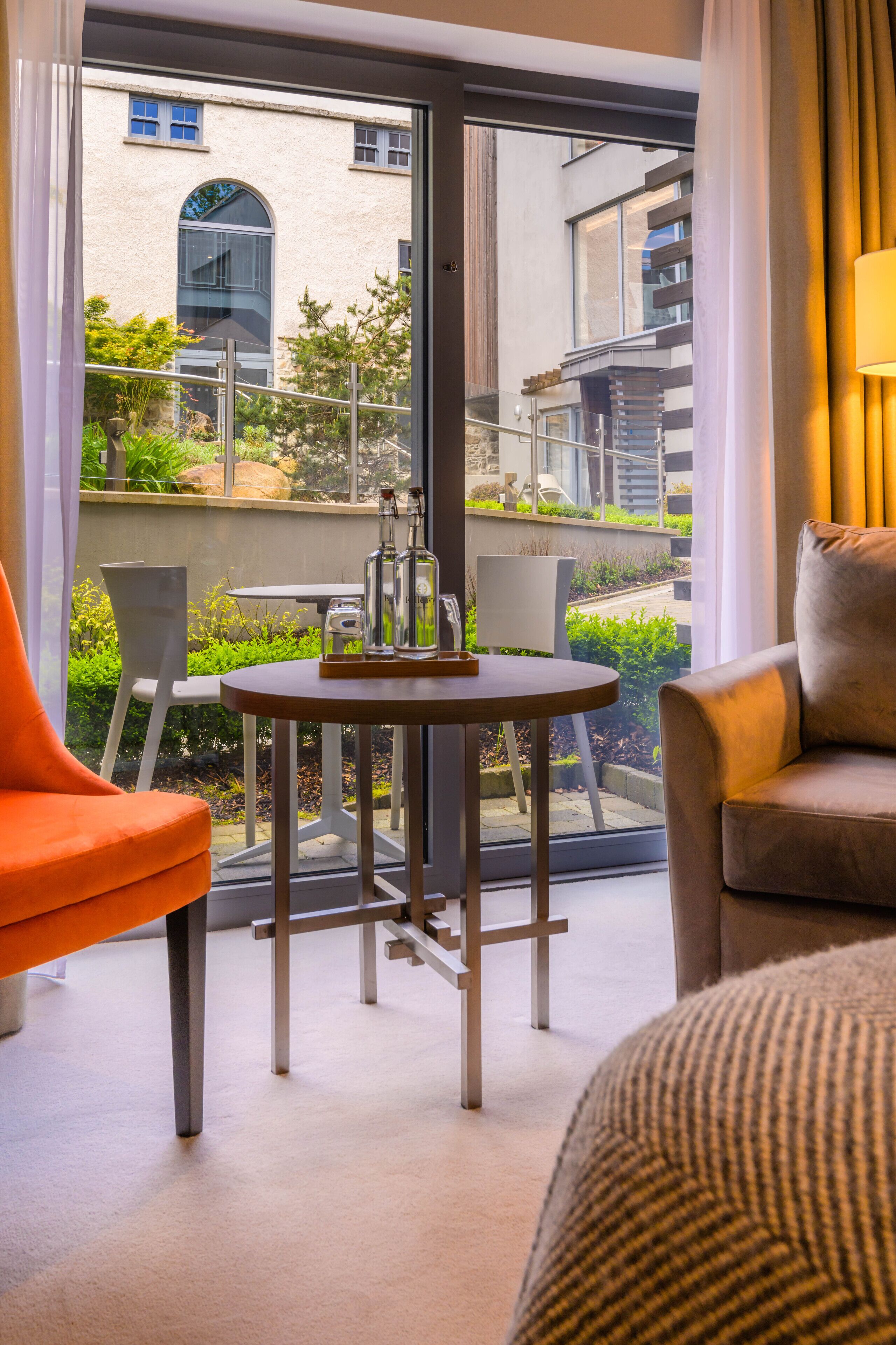 A modern hotel room with a large window, a round table with two bottles, and two chairs.