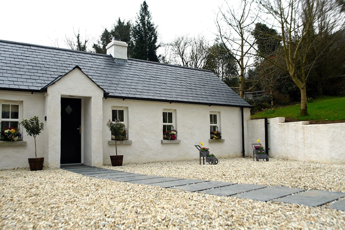 A white cottage with a grey tiled roof, a stone wall to the left, and gravel driveway.
