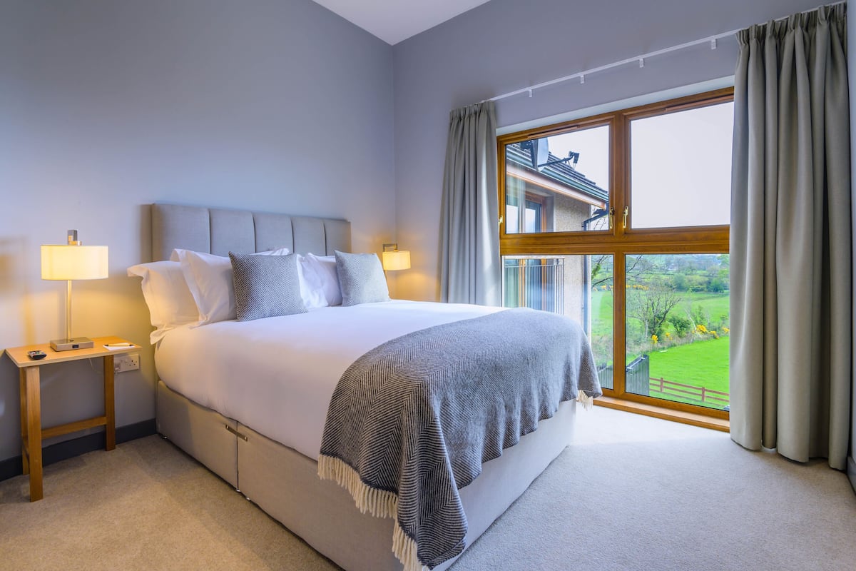 A bedroom with a large bed, two bedside tables with lamps, a view of a green landscape through a window.