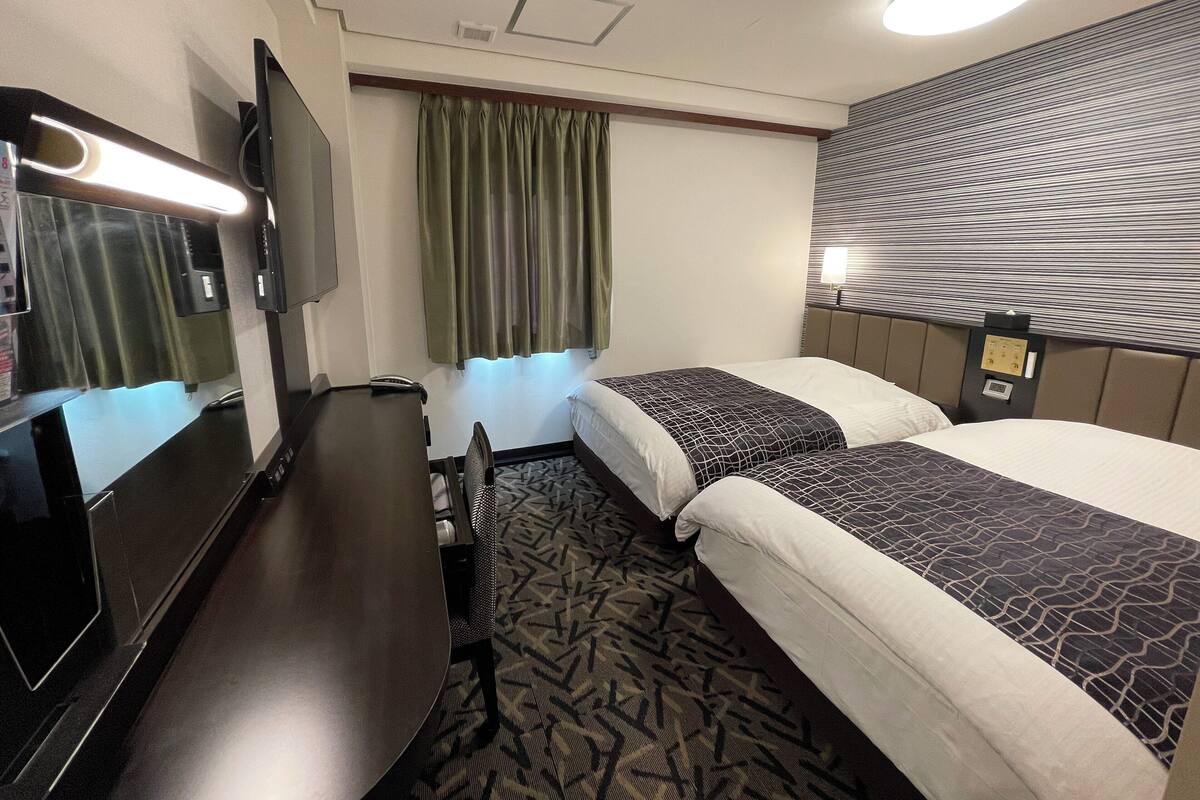 A hotel room with two beds, a desk, a TV, and a window with curtains.