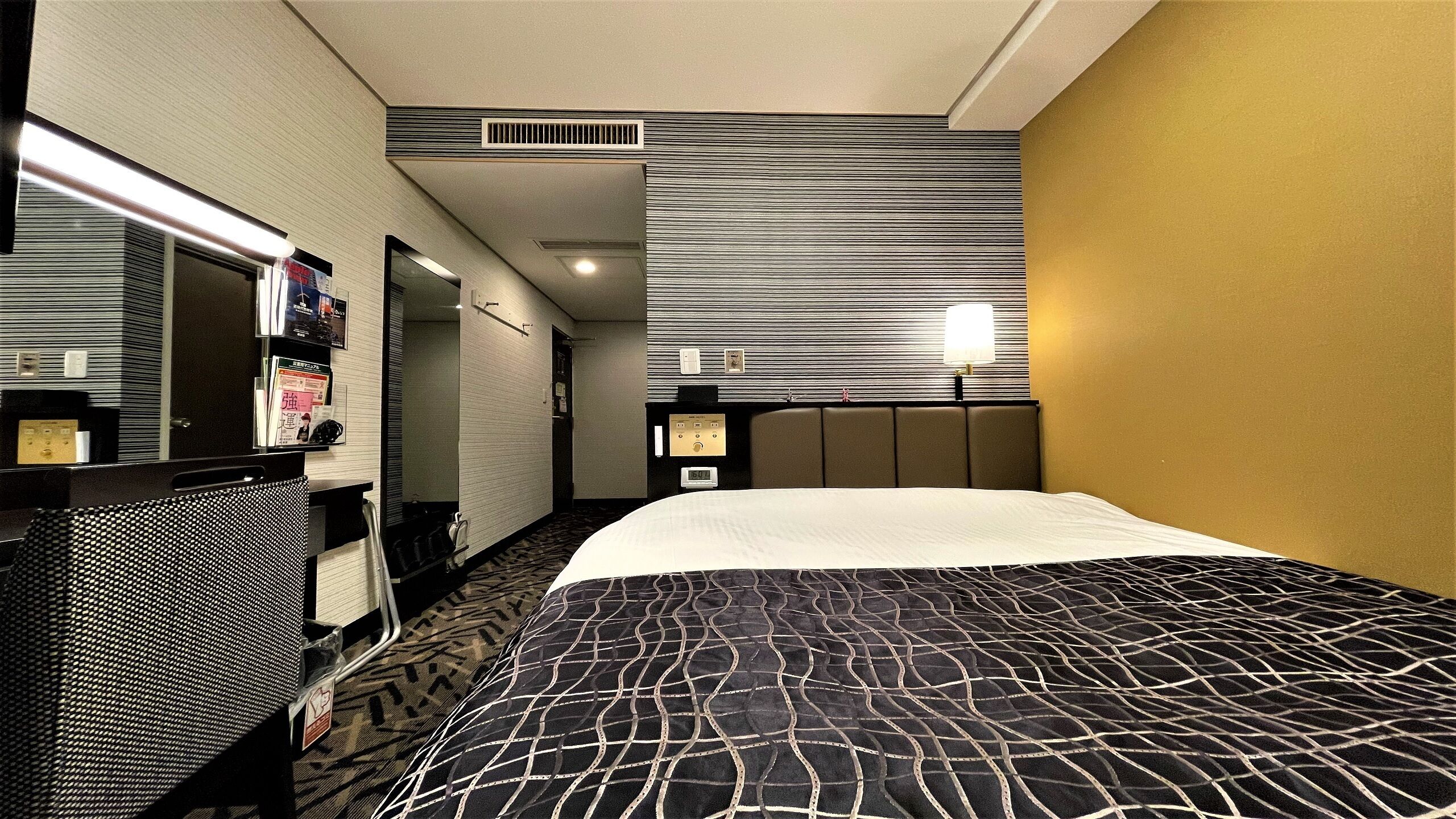 A hotel room with a bed, a desk, a mirror, a wall-mounted TV, and a window.