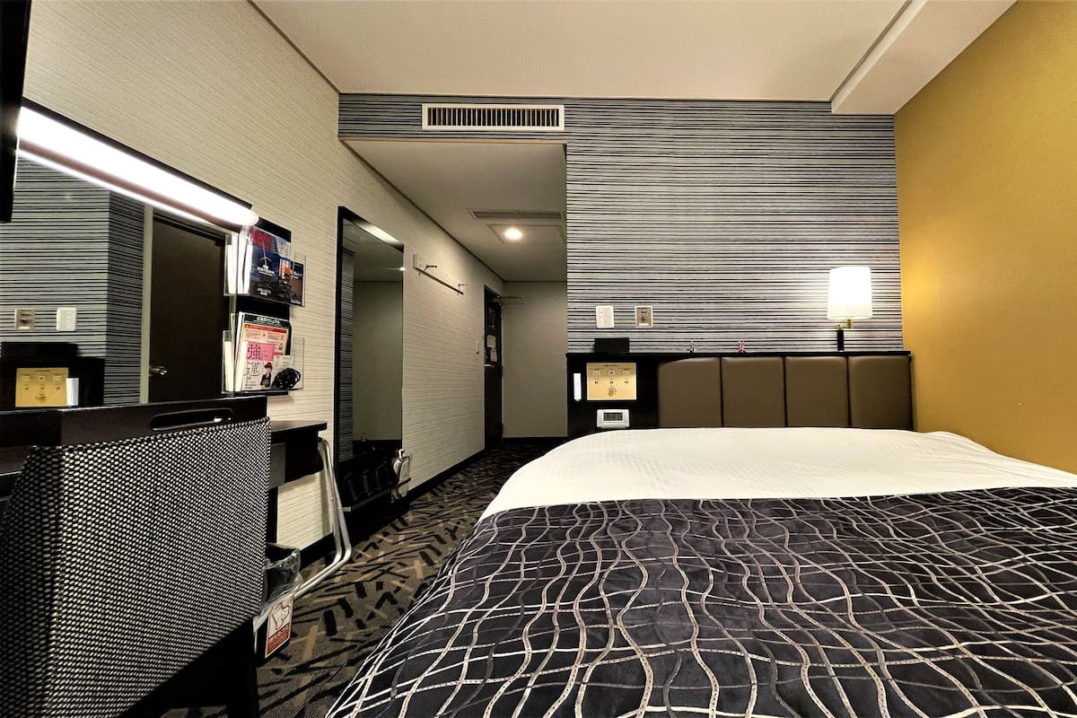 A hotel room with a bed, a desk, a mirror, a wall-mounted TV, and a window.