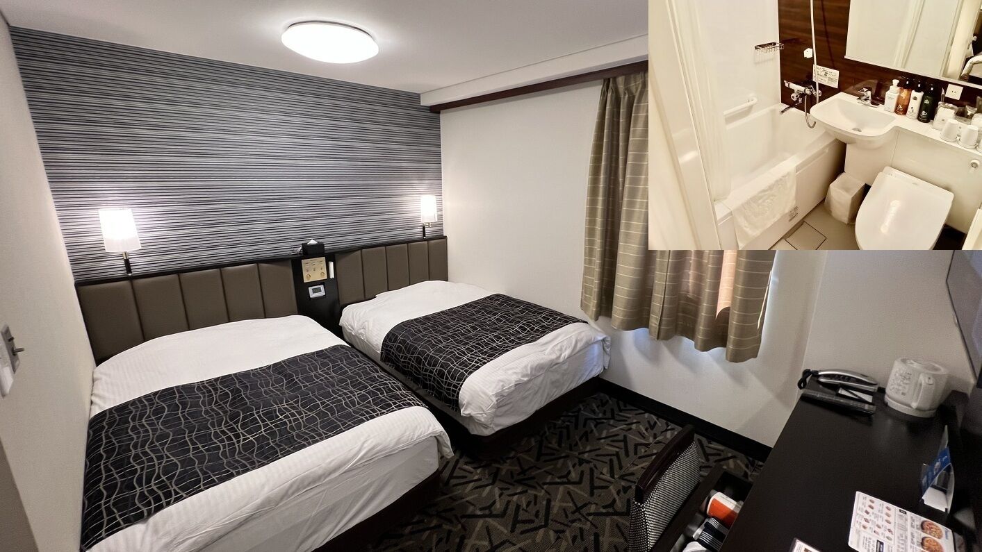 A hotel room with two beds, a headboard, a desk with a chair, a bathroom with a sink and shower, and a patterned carpet.