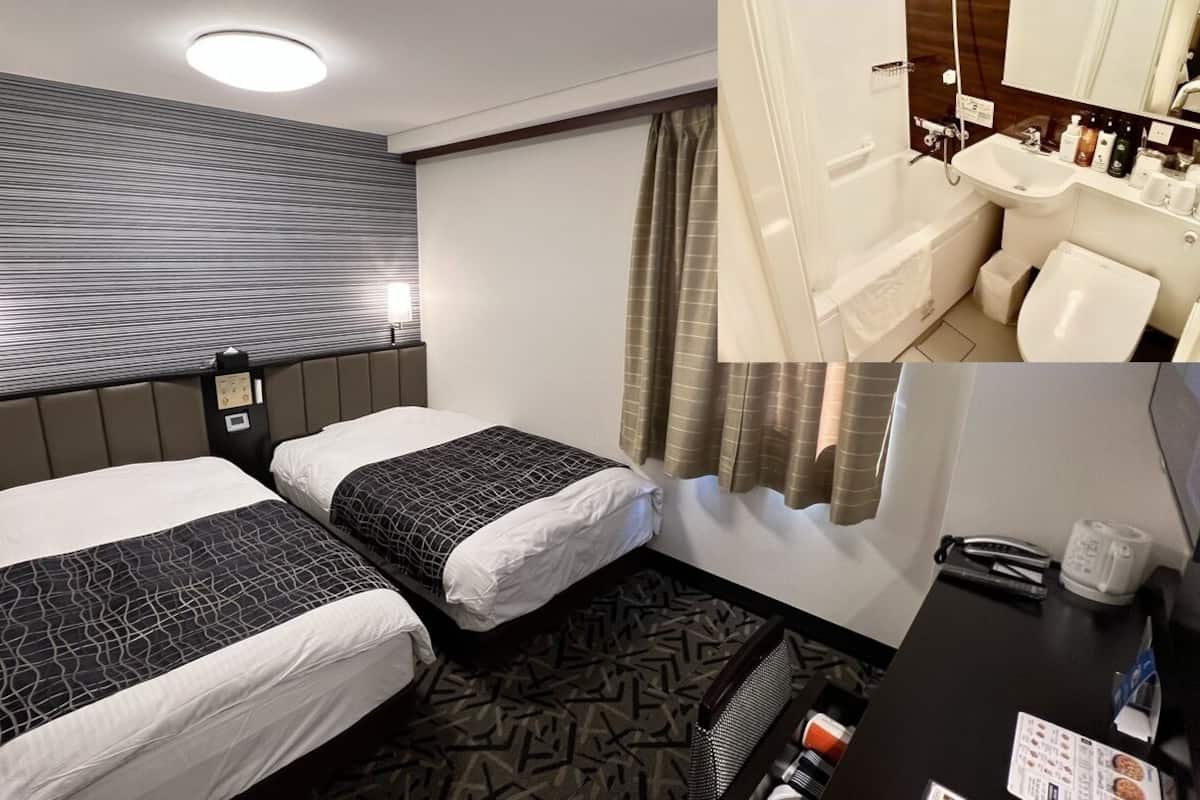 A hotel room with two beds, a headboard, a desk with a chair, a bathroom with a sink and shower, and a patterned carpet.