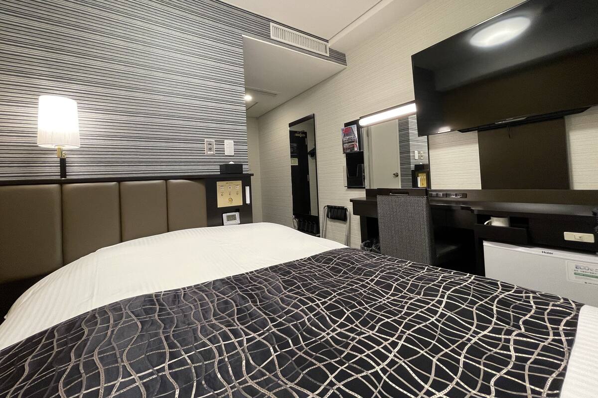A hotel room with a bed, a desk, a chair, a mirror, and a wall-mounted heater.