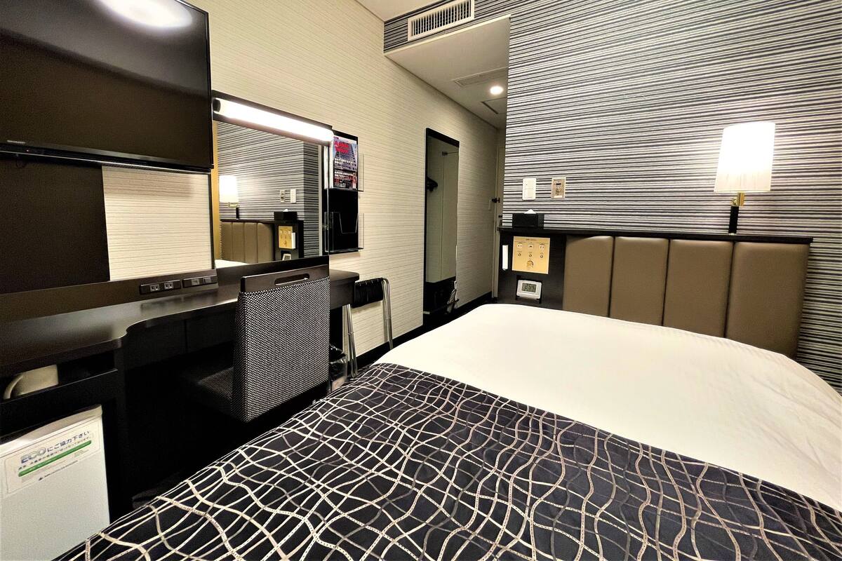 A hotel room with a bed, a desk, a chair, a TV, and a wall-mounted control panel.