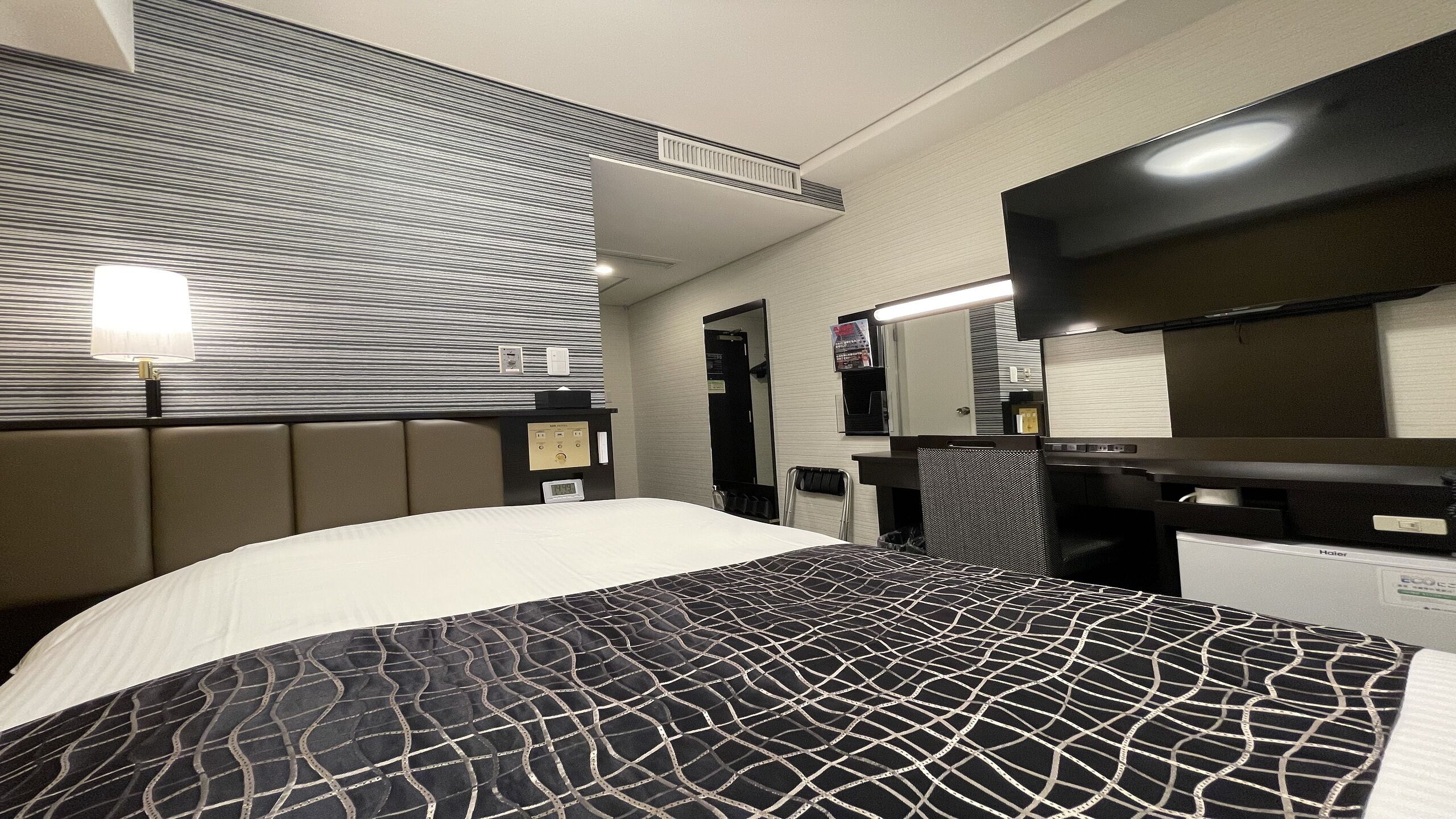 A hotel room with a bed, a desk, a chair, a mirror, and a wall-mounted TV.