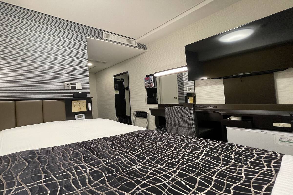 A hotel room with a bed, a desk, a chair, a mirror, and a wall-mounted TV.