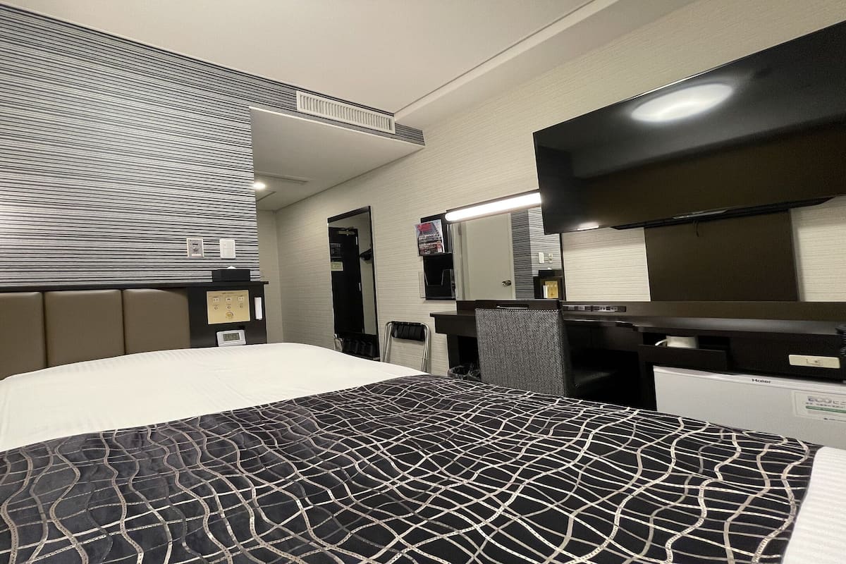 A hotel room with a bed, a desk, a chair, a mirror, and a wall-mounted TV.