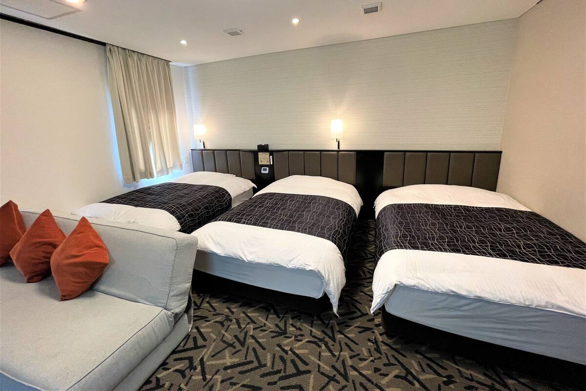 A hotel room with two beds, a sofa, and a wall-mounted lamp.