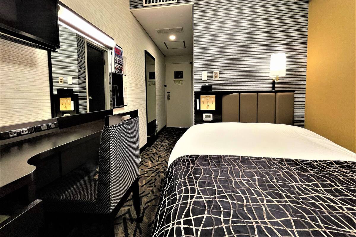 A hotel room with a bed, a desk, a chair, a mirror, a clock, and a door.
