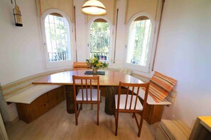 A dining area with a wooden table, four chairs, and a bench. There are three arched windows providing natural light.