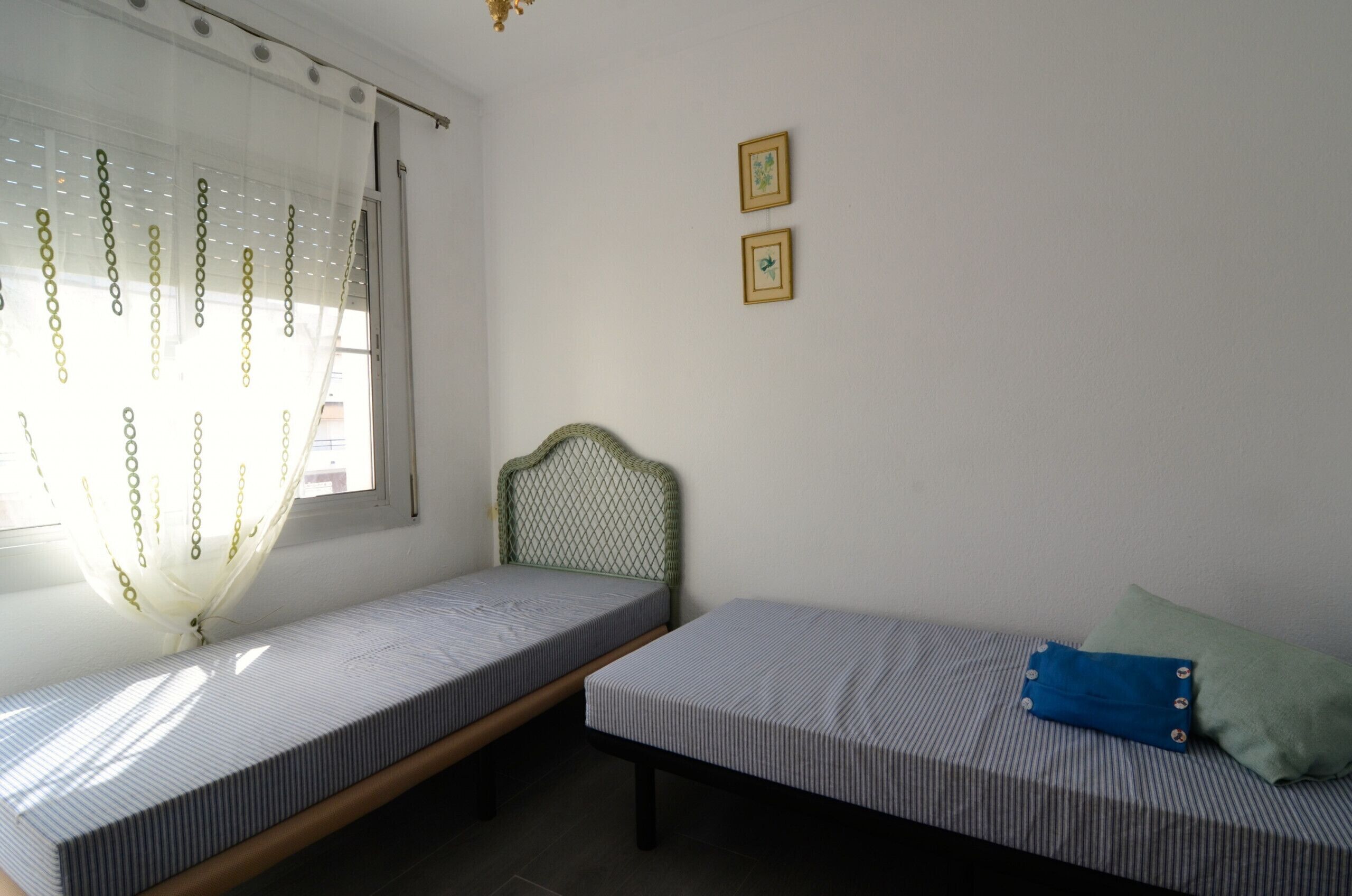 Une chambre avec deux lits simples, une fenêtre avec des rideaux légers et deux tableaux encadrés au mur.