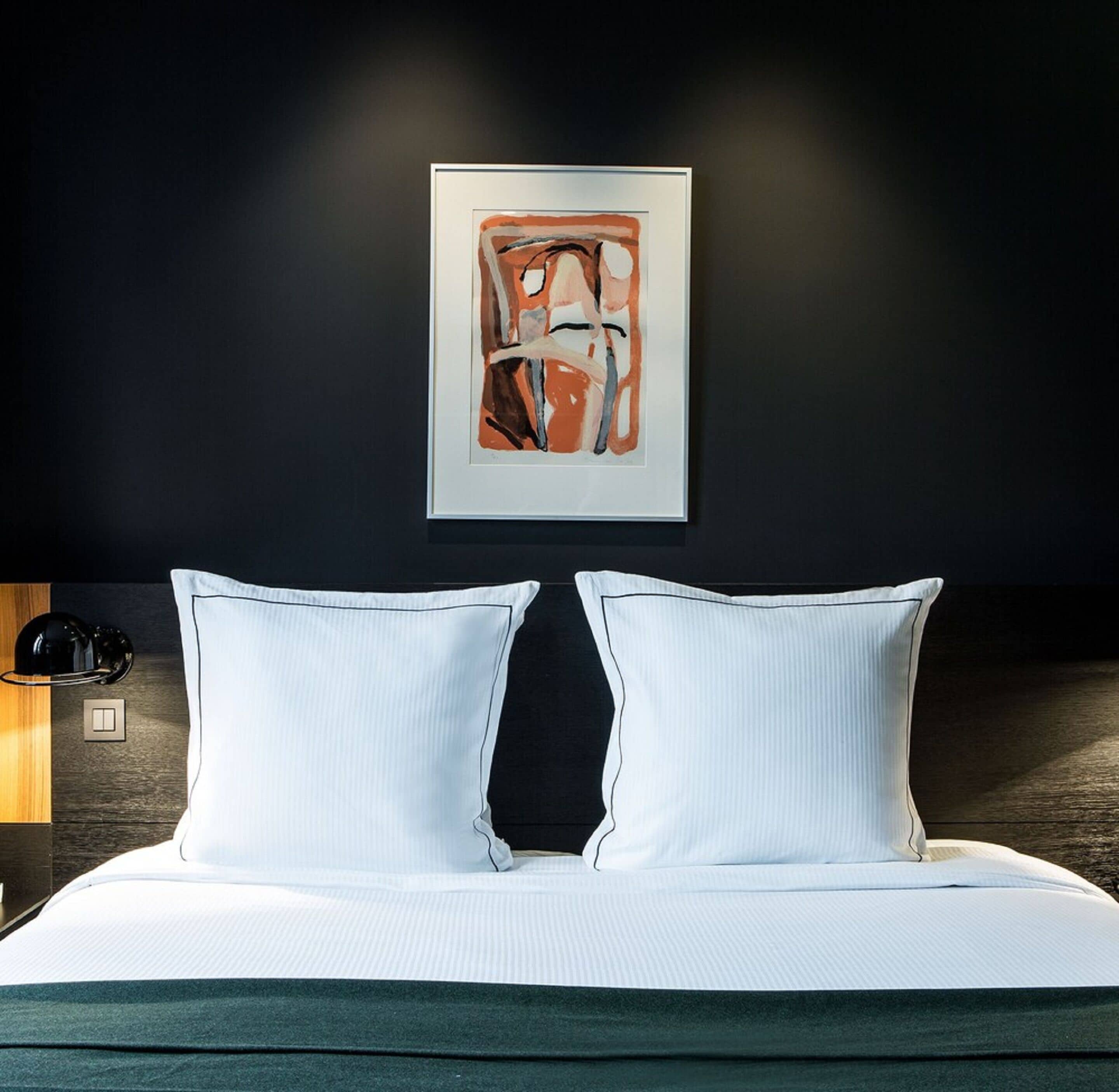 Un letto rifatto con lenzuola bianche e due cuscini, un quadro astratto incorniciato sulla parete e una lampada fissata al muro.