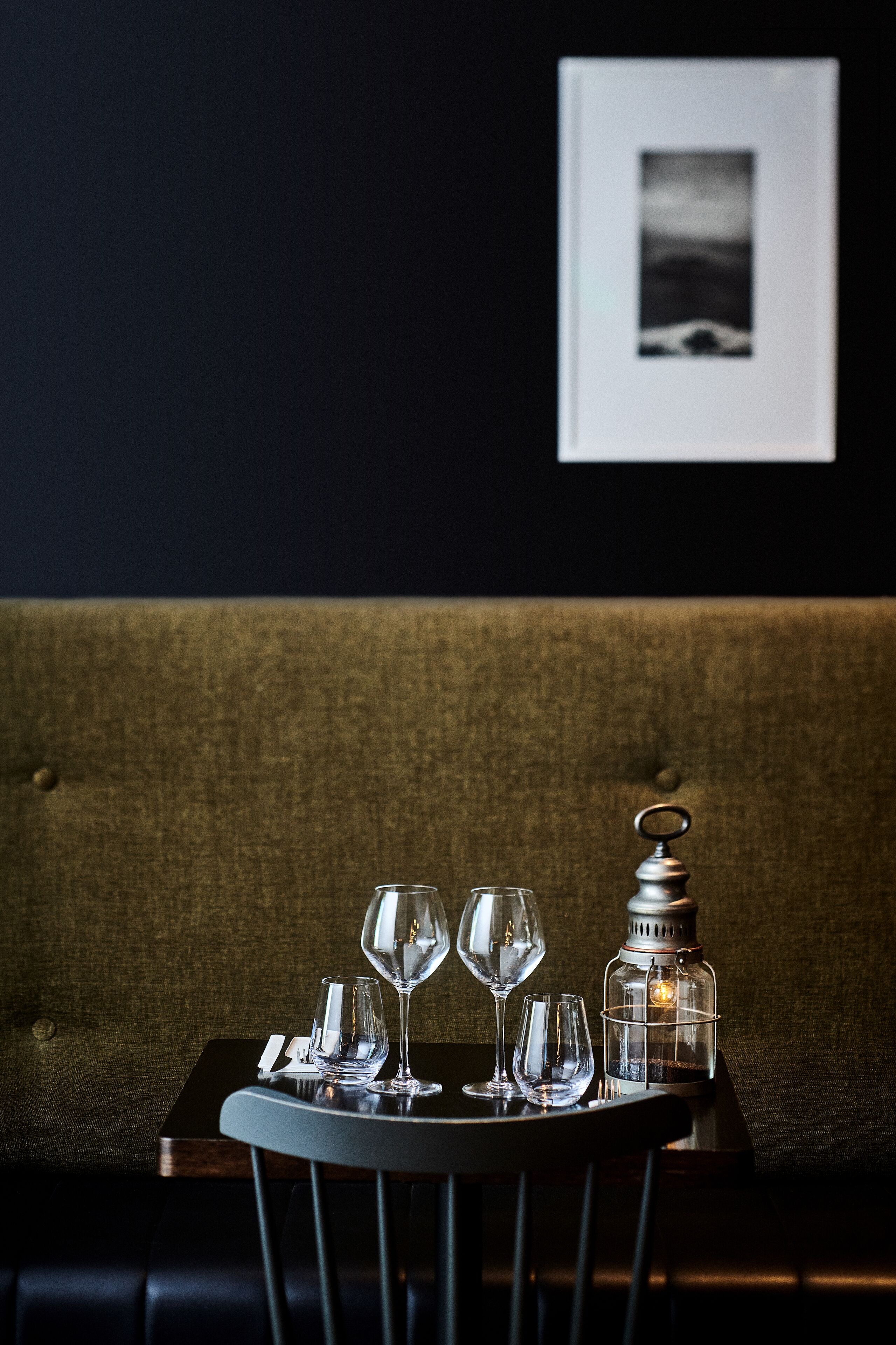A table with three wine glasses and a candle in a dimly lit room with a dark wall and a framed picture.