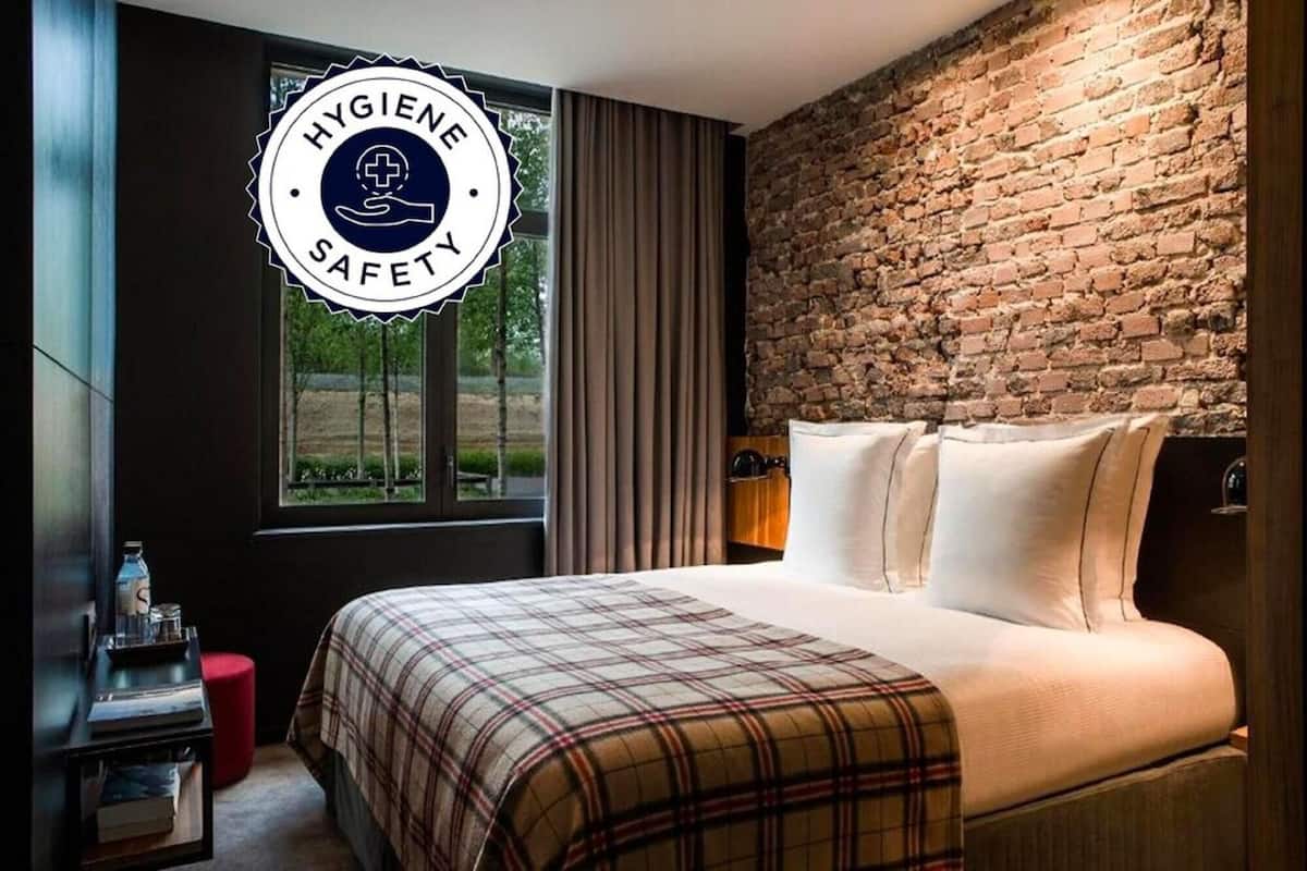 A bedroom with a brick wall, a window with curtains, a bed with a plaid blanket, and a nightstand with a bottle.