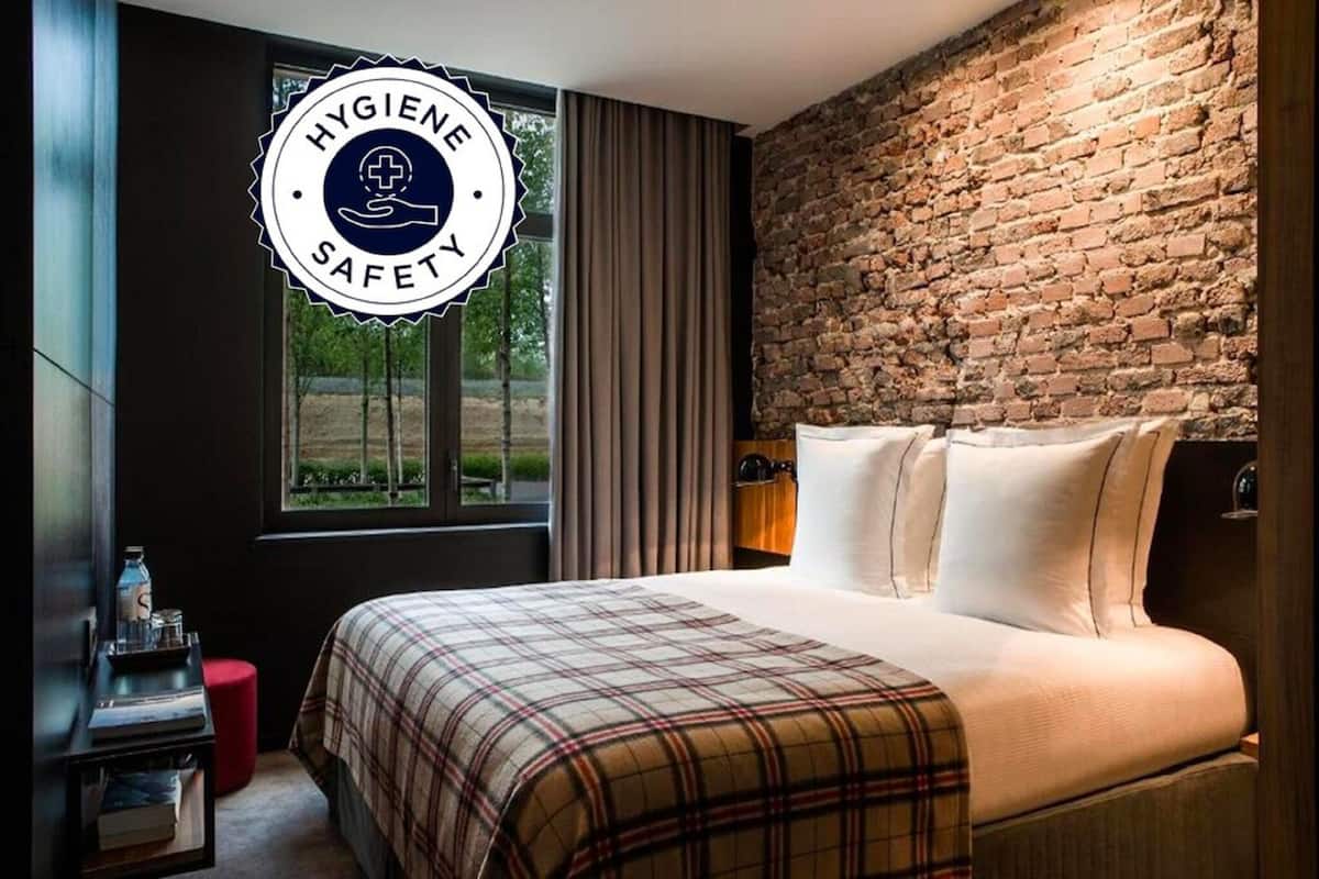 Ein Hotelzimmer mit einem großen Bett, einer Ziegelwand, einem Fenster mit Vorhängen und einem Schild mit Hygiene- und Sicherheitsinformationen.