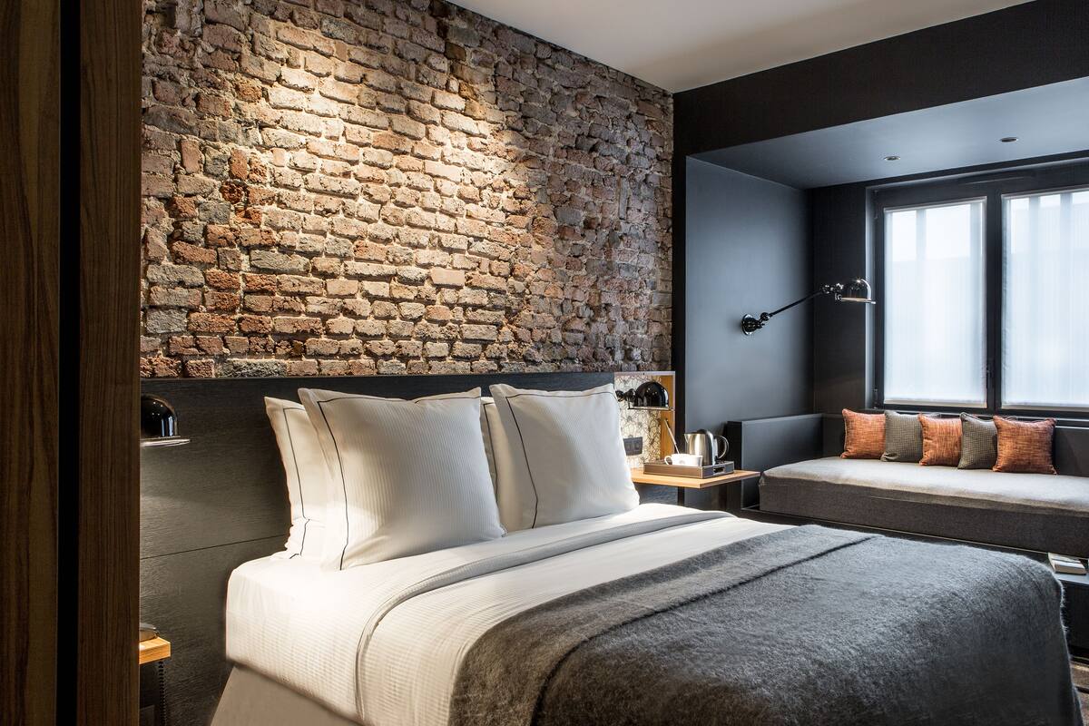 Ein modernes Schlafzimmer mit Backsteinwand, einem großen Bett mit vielen Kissen, einem Nachttisch, einer kleinen Couch mit Kissen und einem Fenster mit Vorhängen.