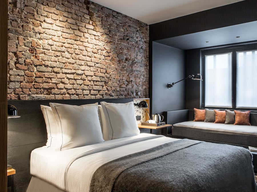 A modern bedroom with a brick wall, a large bed with multiple pillows, a bedside table, a small sofa with cushions, and a window with curtains.