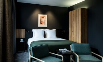 Una habitación de hotel moderna con una cama grande, dos mesitas de noche, un armario de madera, dos sillones tapizados y una obra de arte enmarcada en la pared.
