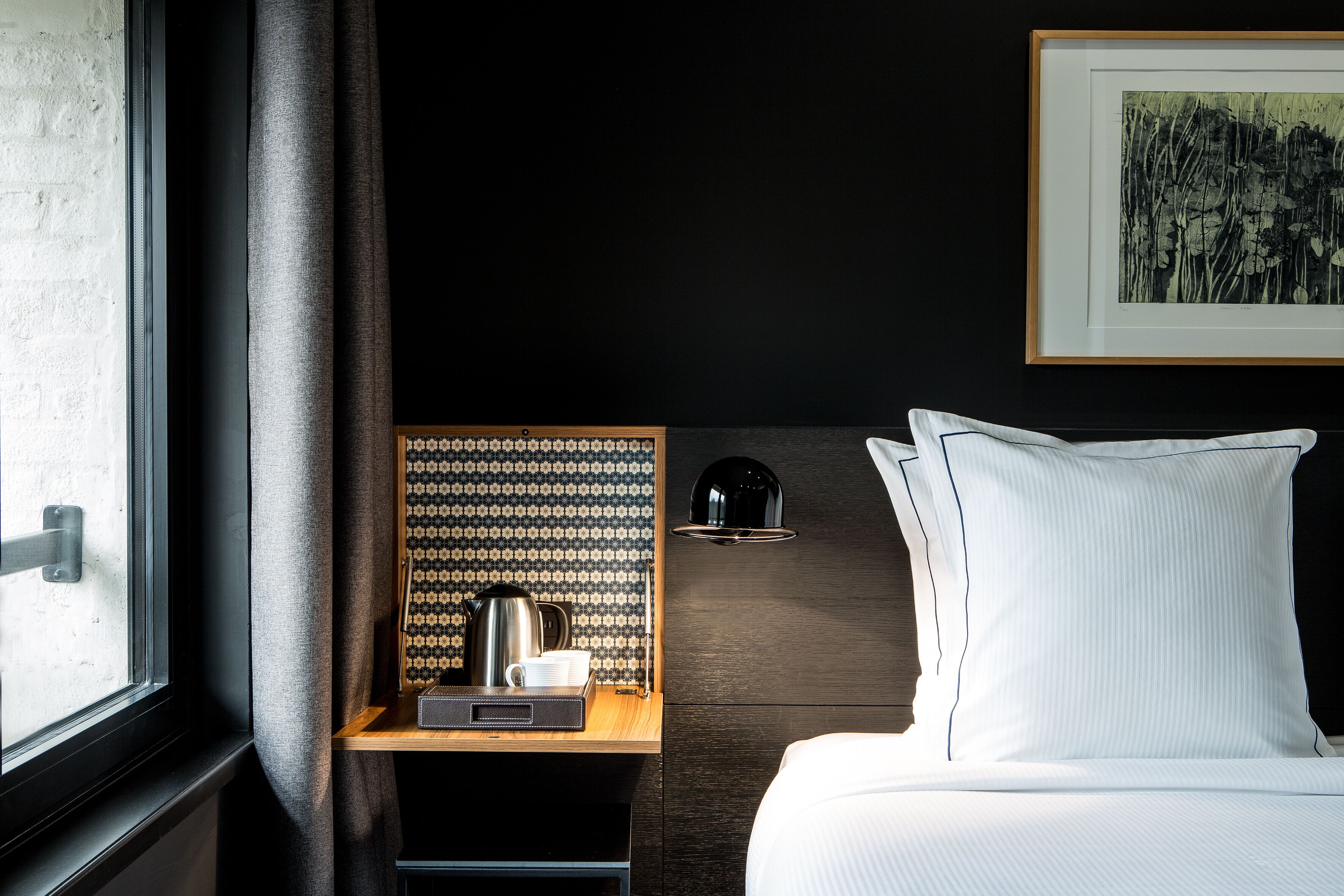 A hotel room with a bed, a nightstand with a coffee maker, a framed picture on the wall, and a window with curtains.