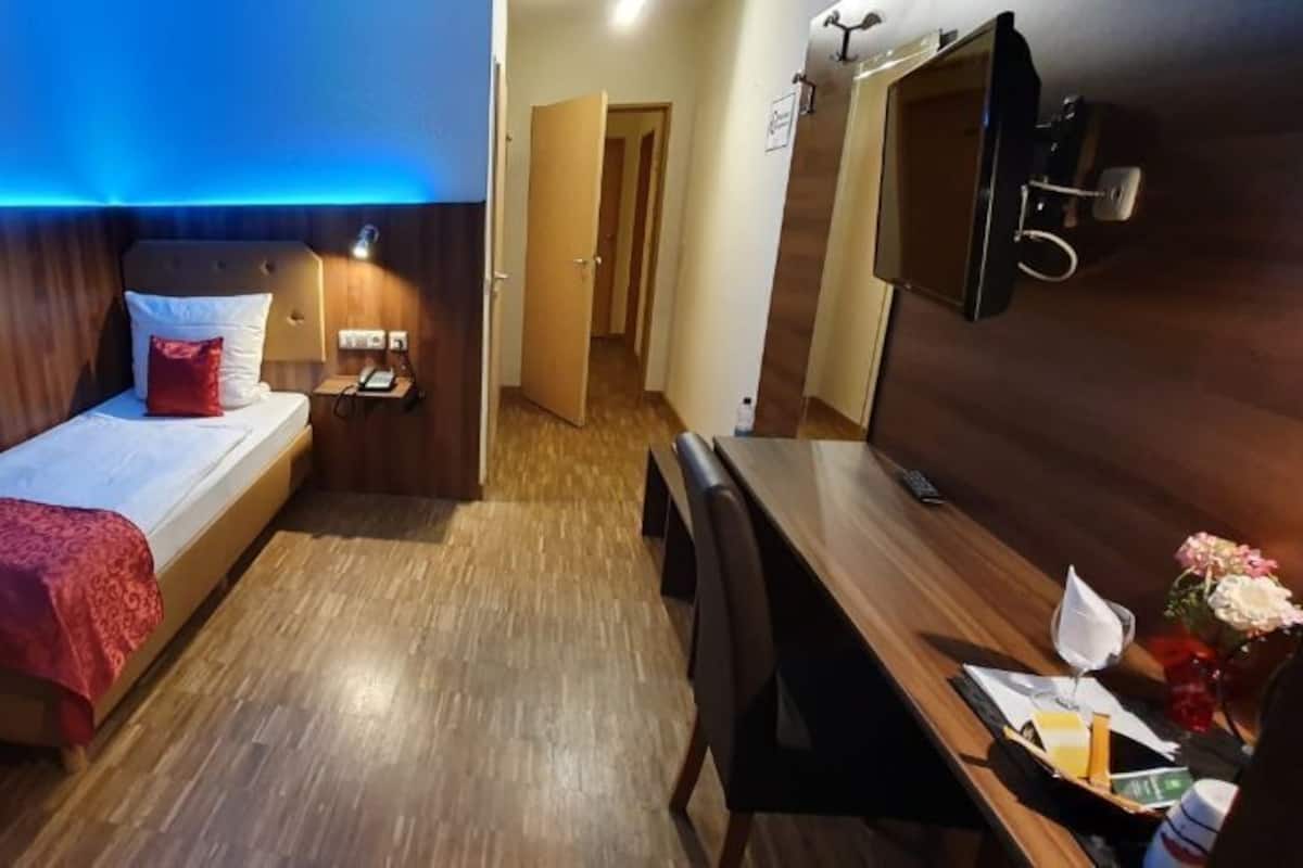 A compact hotel room with a single bed, a small bedside table, a desk with a chair, and a TV mounted on the wall.