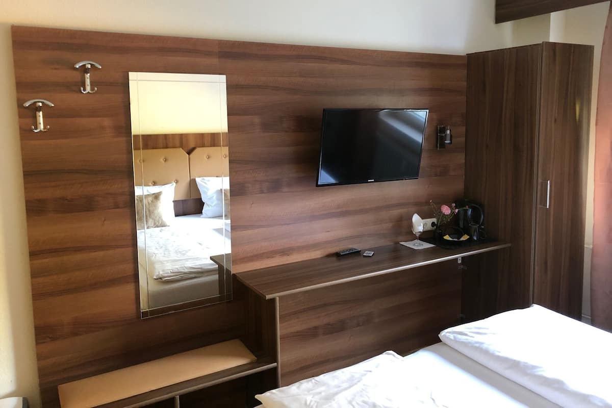 A hotel room with a wooden headboard, a flat-screen TV mounted on the wall, a bedside table with a lamp, and a large wardrobe.