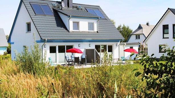 Ein zweistöckiges Haus mit einem geneigten Dach, Solaranlagen und einer überdachten Terrasse mit roten Sonnenschirmen.