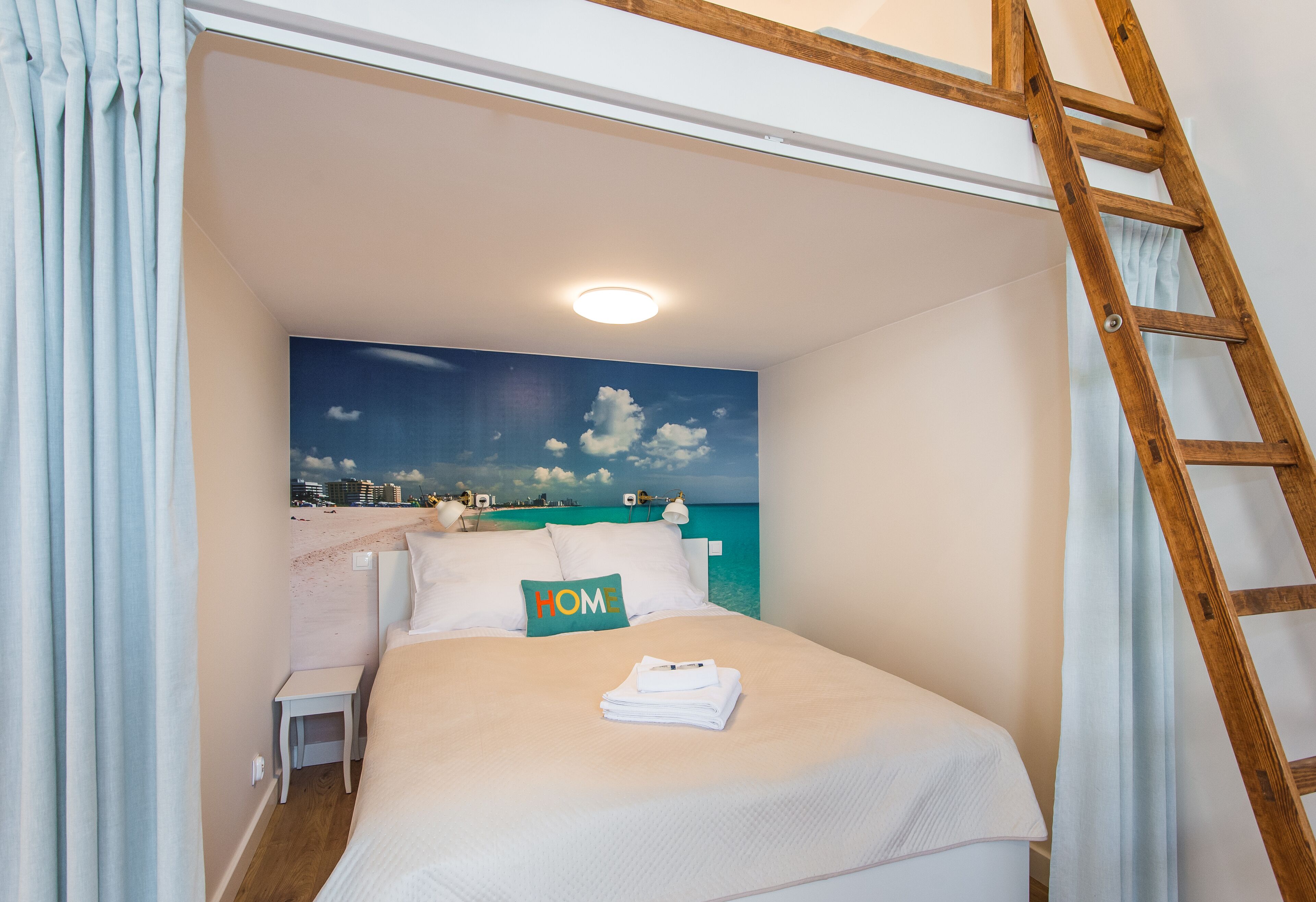 Een net opgemaakt bed met een kussen waarop 'HOME' staat, een opgevouwen handdoek, een houten ladder die tegen de muur leunt, en een schilderij van een strandzicht aan de muur.